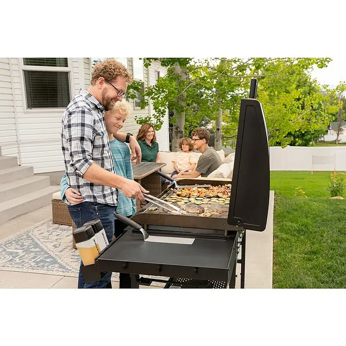 Blackstone Plancha Grill - Superficie de la parrilla (An x Pr): 91 x 55 cm, Negro, Número de quemadores: 4 ud. - Plancha exterior negra utilizada por un hombre y un niño para cocinar verduras y carne para una familia.