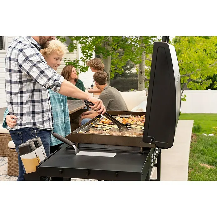 Blackstone Plancha Grill - Superficie de la parrilla (An x Pr): 91 x 55 cm, Negro, Número de quemadores: 4 ud. - Un hombre y un niño cocinan filetes y verduras en una plancha exterior negra en una reunión familiar. La plancha tiene una superficie de cocción grande y oscura.