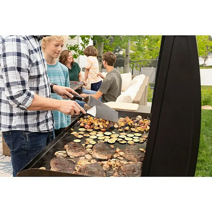 Blackstone Plancha Grill - Superficie de la parrilla (An x Pr): 91 x 55 cm, Negro, Número de quemadores: 4 ud. - Hombre usa espátulas de parrilla de metal con mango negro para cocinar bistec, verduras y champiñones en una plancha exterior para una comida familiar.