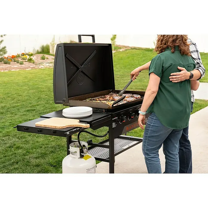 Blackstone Plancha Grill - Superficie de la parrilla (An x Pr): 91 x 55 cm, Negro, Número de quemadores: 4 ud. - Una plancha Blackstone negra cocinando filetes y verduras al aire libre, con dos personas, un tanque de propano y estantes laterales.
