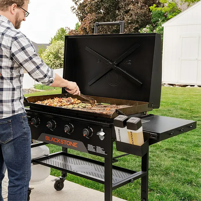 Blackstone Plancha Grill - Superficie de la parrilla (An x Pr): 91 x 55 cm, Negro, Número de quemadores: 4 ud. - Un hombre cocina al aire libre en una gran plancha Blackstone negra, mostrando su amplia superficie de cocción y funciones.