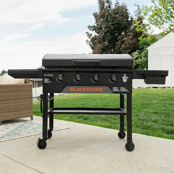 Blackstone Plancha Grill - Superficie de la parrilla (An x Pr): 91 x 55 cm, Negro, Número de quemadores: 4 ud. - Plancha Blackstone negra con cuatro mandos de control en un patio. Estructura de metal negro y superficie de cocción.