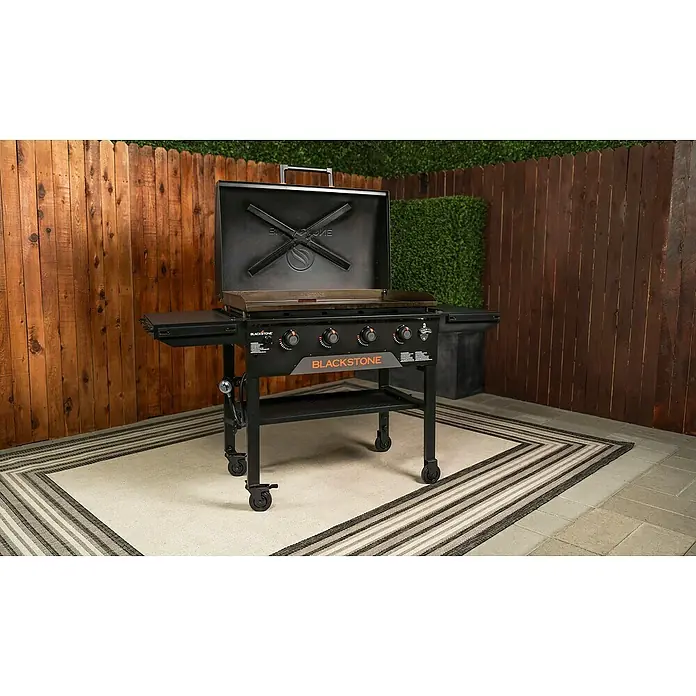 Blackstone Plancha Grill - Superficie de la parrilla (An x Pr): 91 x 55 cm, Negro, Número de quemadores: 4 ud. - Plancha Blackstone negra con ruedas y tapa abierta, mostrando la superficie de cocción y los mandos de control, sobre una alfombra de patio.