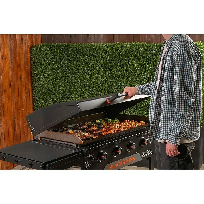Blackstone Plancha Grill - Superficie de la parrilla (An x Pr): 91 x 55 cm, Negro, Número de quemadores: 4 ud. - Una persona abre la tapa negra de una plancha Blackstone, cocinando carne, patatas y brócoli.