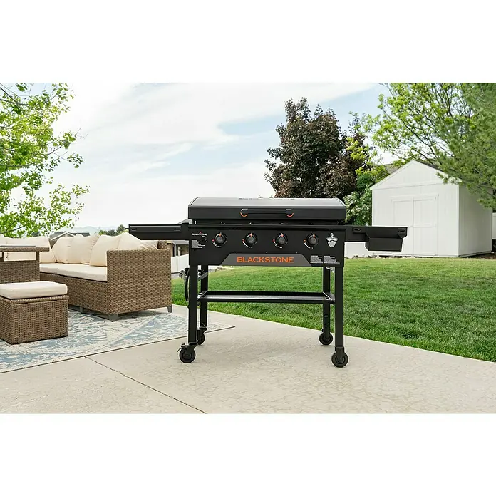 Blackstone Plancha Grill - Superficie de la parrilla (An x Pr): 91 x 55 cm, Negro, Número de quemadores: 4 ud. - Plancha Blackstone negra con detalles naranjas en un patio junto a muebles de exterior.