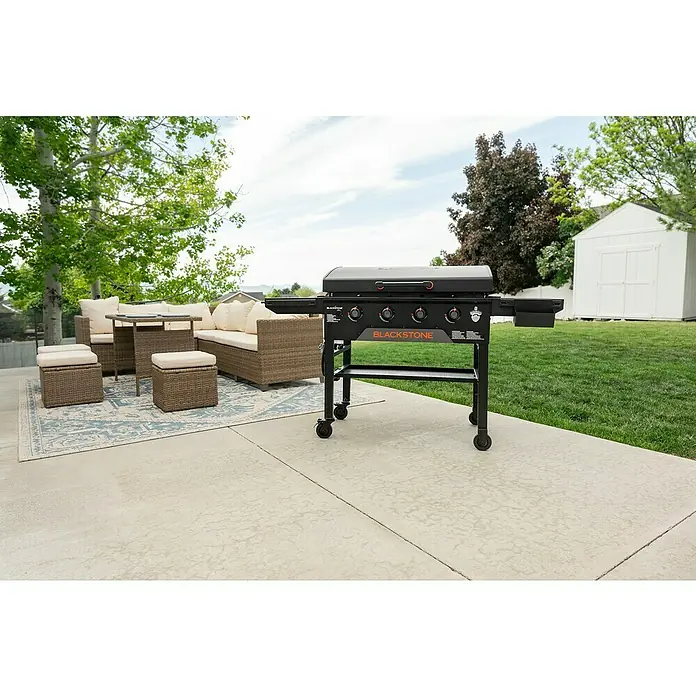 Blackstone Plancha Grill - Superficie de la parrilla (An x Pr): 91 x 55 cm, Negro, Número de quemadores: 4 ud. - Plancha Blackstone negra con detalles naranjas en un patio, lista para cocinar al aire libre junto a muebles de mimbre.