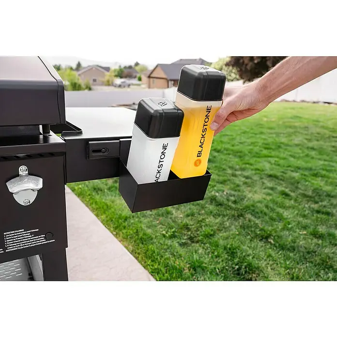 Blackstone Plancha Grill - Superficie de la parrilla (An x Pr): 91 x 55 cm, Negro, Número de quemadores: 4 ud. - Una mano coloca dos botellas Blackstone (blanca, amarilla) en un soporte negro unido a una parrilla negra exterior.