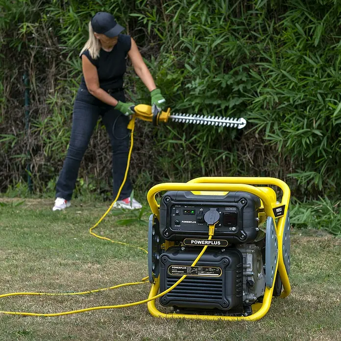 PowerPlus Generador POWX5230 - 3 kW, Volumen del depósito: 10 l - Un generador inversor amarillo POWERPLUS alimenta un cortasetos usado por una mujer para podar un seto.