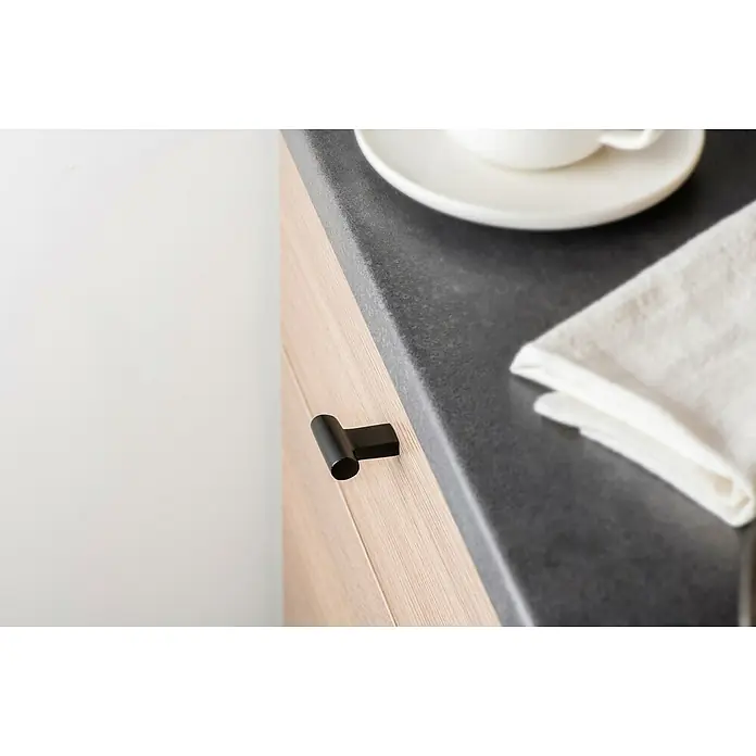 Rei Möbelknopf - Ø x L: 12 x 33 mm, Schwarz - Schwarzer zylindrischer Schrankgriff an einem hellen Holzschrank neben einer dunkelgrauen Arbeitsplatte.
