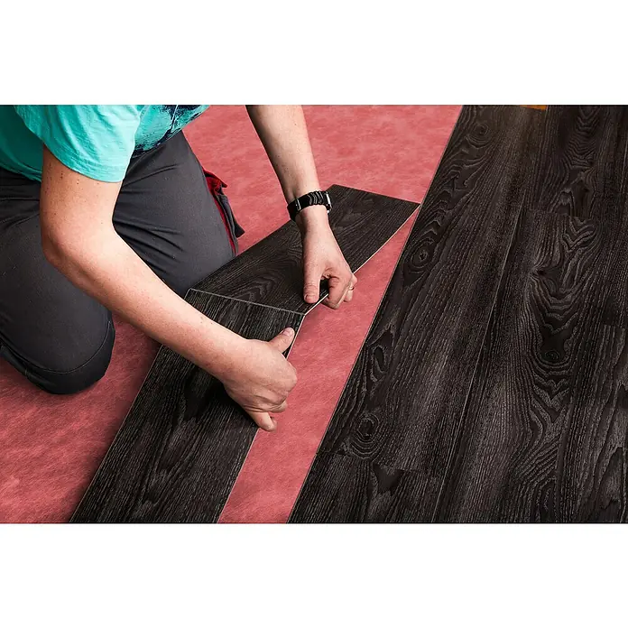 b!design Unterlagsmatte - 10 m², Stärke: 1,4 mm - Person verlegt dunkle Laminatdielen in Holzoptik auf einer roten Unterlage.