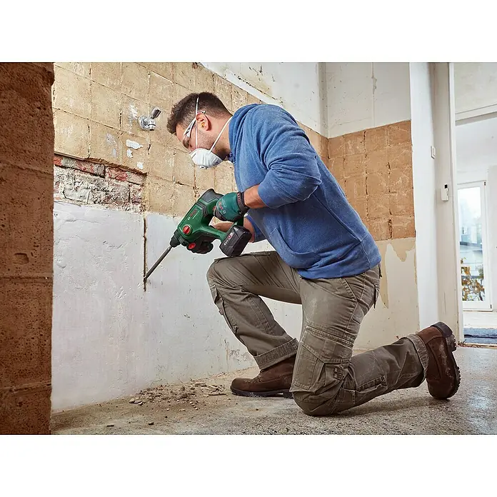 Bosch Professional AMPShare 18V Akku-Kombihammer UniversalHammer 18V - 18 V, 1 Akku, 4 Ah, 2 J - Person in Schutzausrüstung benutzt einen grün-schwarzen Bosch Bohrhammer, um eine Wand zu bearbeiten.