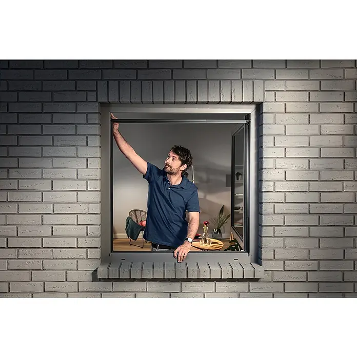 Tesa Insektenschutzfenster FALT für Fenster - Weiß, Montage Insektengitter: Klemmen - Mann justiert ein dunkles, ausziehbares Fensterrollo in einem Fenster einer Ziegelwand.