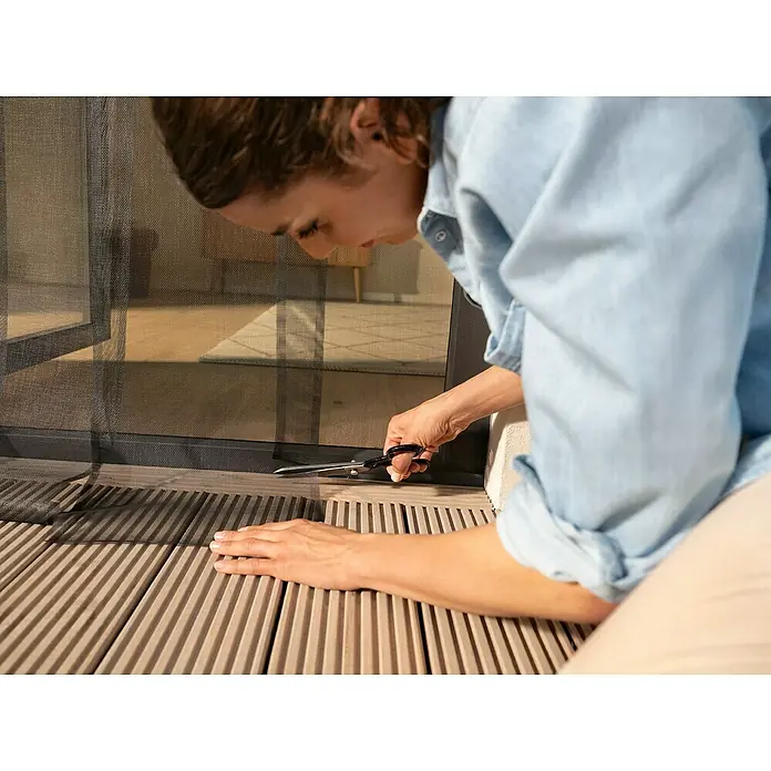 Tesa Insect Stop Insektenschutzvorhang Lamellenvorhang - B x H: 1,2 x 2,5 m, Montage Insektengitter: Ankletten - Person schneidet ein dunkles Fliegengittergewebe zur Anpassung für Tür oder Fenster.