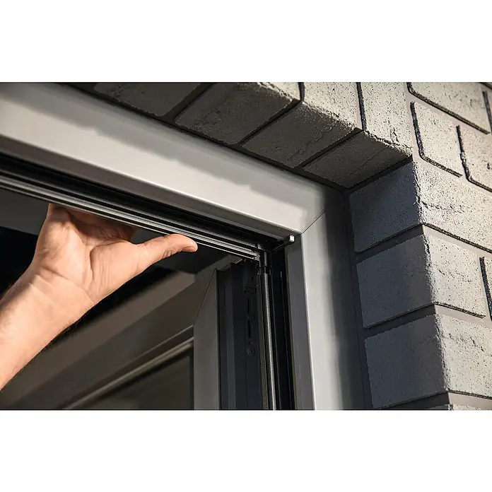 Tesa Insektenschutzfenster FALT für Fenster - Weiß, Montage Insektengitter: Klemmen - Hand justiert einen dunkelgrauen Fensterrahmen, eingesetzt in eine graue Ziegelwand.