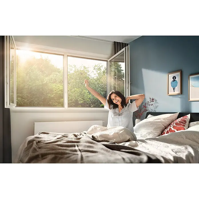 Tesa Insect Stop Pollenschutzgitter Pollen Protection - B x H: 130 x 150 cm, Montage Insektengitter: Ankletten, Anthrazit - Frau streckt sich im Bett in einem hellen Schlafzimmer mit geöffnetem Fenster.