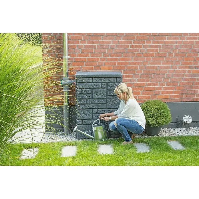 Arves Regenspeicher Maurano - 300 l, Black Granitfarben, 1 Stk. - Frau befüllt eine Gießkanne an einem grauen Regenspeicher mit Steinmuster vor einer roten Backsteinmauer in einem grünen Garten.