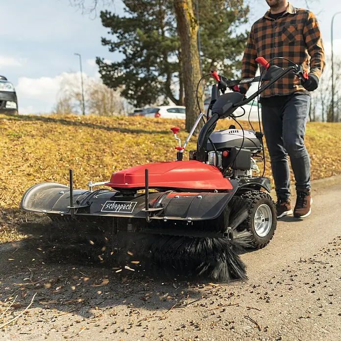 Scheppach Kehrmaschine SC2200PE 3 in 1 - 3,6 kW, Arbeitsbreite: 100 cm, 1 x SCHEPPACH Benzin Kehrmaschine - Eine Person bedient eine rot-schwarze Kehrmaschine, die Blätter und Schmutz von einem gepflasterten Weg entfernt.