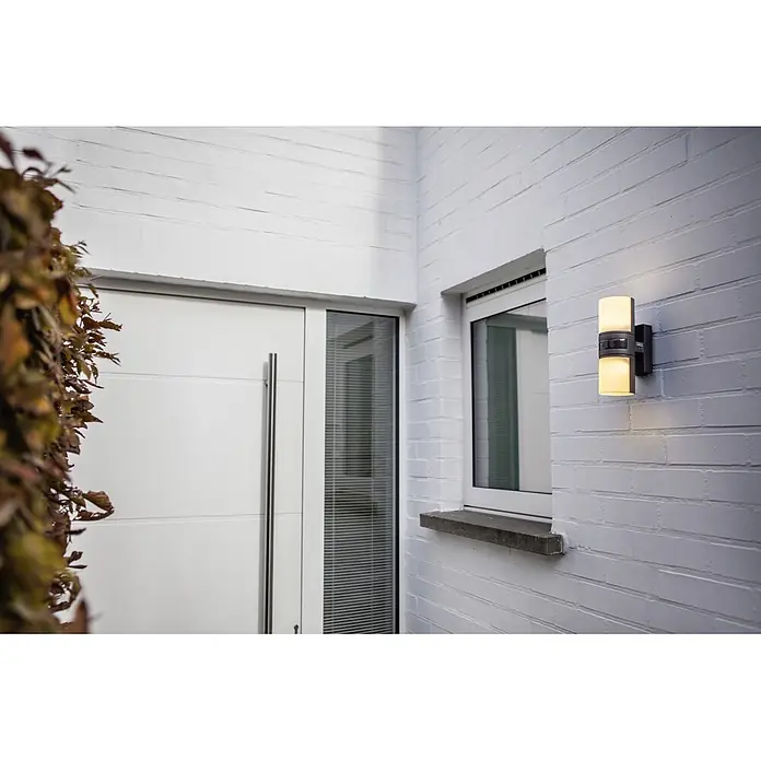 Lutec LED-Sensor-Außenwandleuchte Cyra - 16 W, L x B x H: 7,8 x 12 x 24,5 cm, Anthrazit/Weiß, Warmweiß - Nahaufnahme einer modernen Außenleuchte mit zwei Lampen, die an einer weißen Backsteinmauer neben einem Fenster angebracht ist.