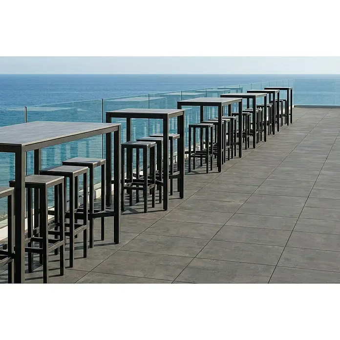 Granulati Zandobbio Terrassenfliese Urban Pro - 60 x 60 x 2 cm, Anthrazit, Matt - Dunkelgraue Outdoor-Bartische und Hocker auf einer Terrasse mit Meerblick.