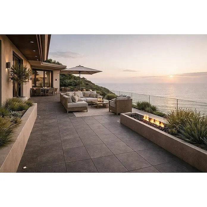Terrassenfliese Urban Pro - 60 x 60 x 2 cm, Anthrazit, Matt - Moderne Außenterrasse mit hellen Loungemöbeln, rechteckiger Stein-Feuerstelle und Essbereich auf dunklen Fliesen mit Meerblick.