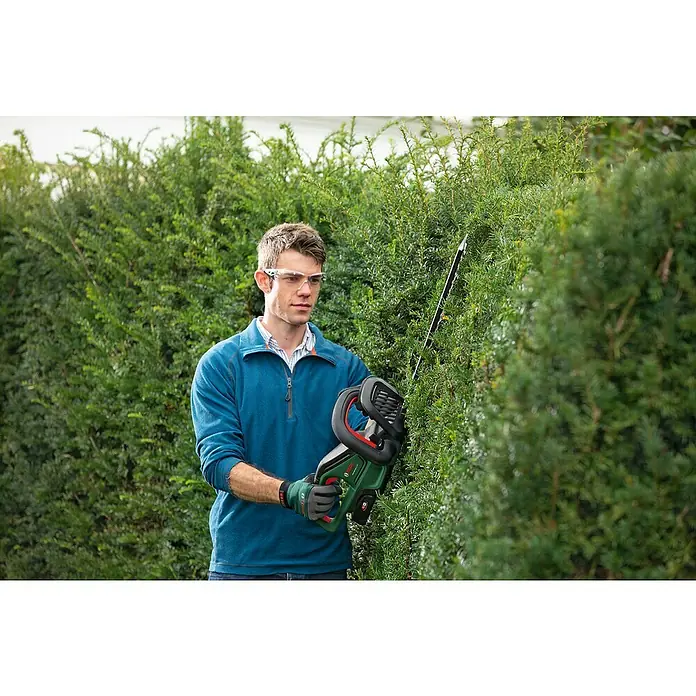 Bosch Power for All 18V Akku-Heckenschere UniversalHedgeCut 18V-50 - 2,5 V, Li-Ionen, Ohne Akku, Schnittlänge: 50 cm - Mann in blauem Oberteil und Schutzbrille schneidet eine grüne Hecke mit einem grün-schwarzen Akku-Heckenschneider.