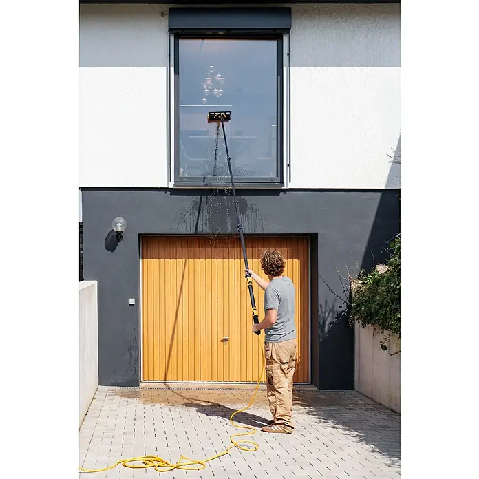 Neptun PV-  & Fassaden Reinigungsset mit verschiedenen Aufsätzen - Teleskopstiel, 120 cm - 490 cm - Mann reinigt mit einer schwarz-gelben, langen, wasserführenden Bürste ein hohes Fenster, während Wasser spritzt.