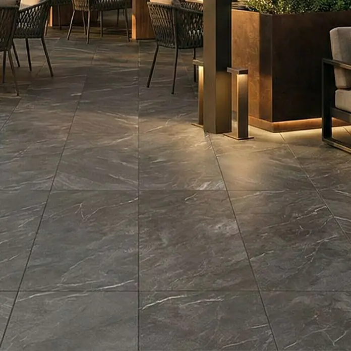 Terrassenfliese Total Stone - 60 x 90 x 2 cm, Grafit, Matt - Dunkelgraue Bodenfliesen in Marmoroptik mit weißen Äderungen, verlegt in einem Geschäftsbereich mit Sitzgelegenheiten und beleuchteten Pflanzgefäßen.