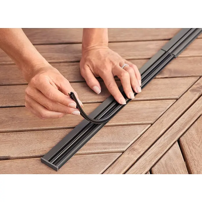 Tesa Insect Stop Lamellenvorhang - B x H: 95 x 220 cm - Hände setzen eine flexible schwarze Gummidichtung in eine schwarze Montageschiene auf Holzoberfläche ein.