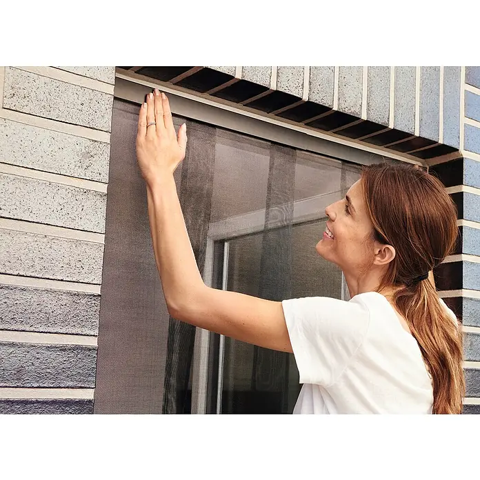 Tesa Insect Stop Lamellenvorhang - B x H: 95 x 220 cm - Frau befestigt dunkles Insektenschutzgitter am Fenster.