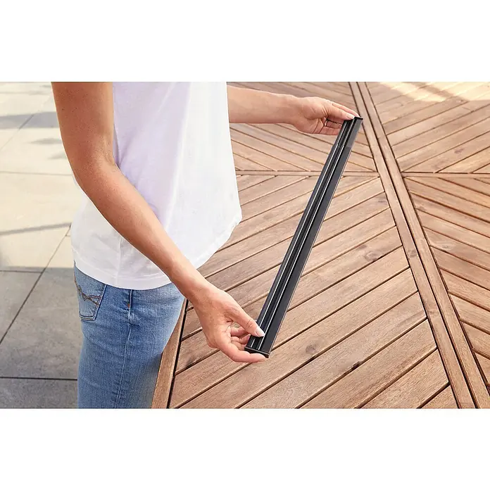 Tesa Insect Stop Lamellenvorhang - B x H: 95 x 220 cm - Eine Person hält ein langes, schwarzes Profil und passt es in eine Holzlatten-Terrasse ein.