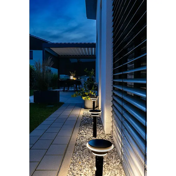 Osram Außenstehleuchte Endura Style solar -  - Zwei schwarze Wegeleuchten mit weißen Ringen beleuchten Kies und Hauswand bei Dämmerung, neben einem gepflasterten Weg zur Terrasse.