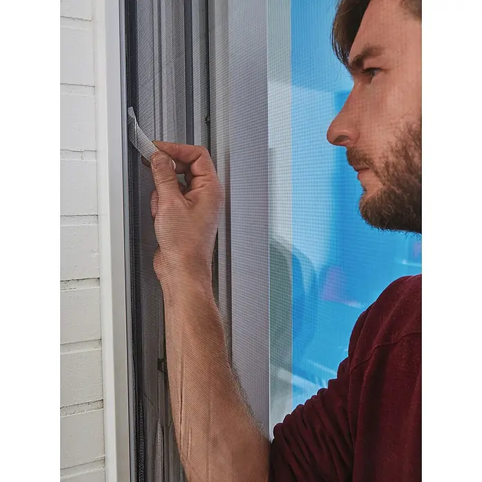 Tesa Insect Stop Insektenschutzgitter Premium - B x H: 120 x 250 cm, Weiß, Montage Insektengitter: Ankletten - Eine Hand justiert einen grauen Plissee-Insektenschutz mit weißem Zugband in einem weißen Fensterrahmen.