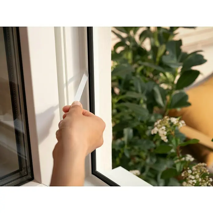 Tesa Insect Stop Insektenschutzgitter Premium - B x H: 100 x 100 cm, Anthrazit, Montage Insektengitter: Ankletten - Eine Hand zieht einen weißen Klebe- oder Klettstreifen von einem weißen Fensterrahmen ab, der eine schwarze Dichtung freilegt.