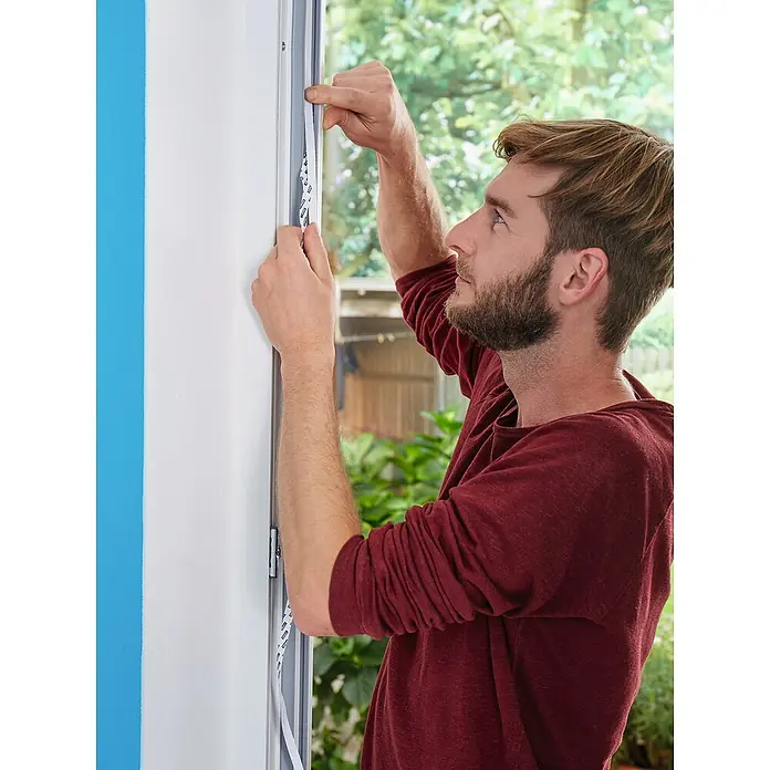 Tesa Insect Stop Insektenschutzgitter Premium - B x H: 120 x 250 cm, Weiß, Montage Insektengitter: Ankletten - Ein Mann bringt weißes Dichtungsband mit blauem Aufdruck an einem Fensterrahmen an.