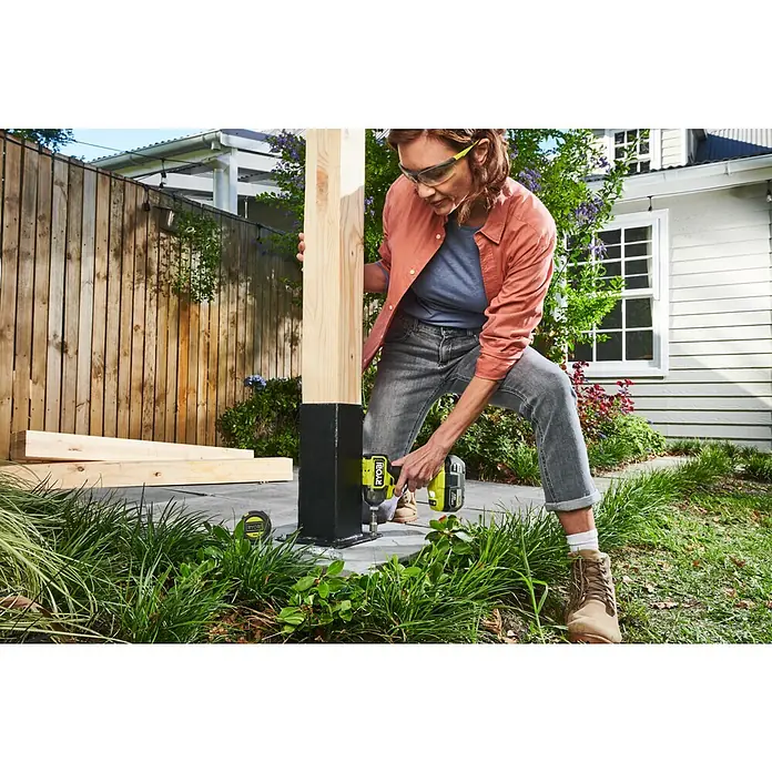 Ryobi ONE+HP Akku-Schlagschrauber RID18X1-0 - 18 V, Ohne Akku, 320 Nm - Frau befestigt Holzpfosten mit gelb-schwarzem Ryobi Akkuschrauber an Metallbasis. Das Werkzeug treibt einen Bolzen in die Basis.