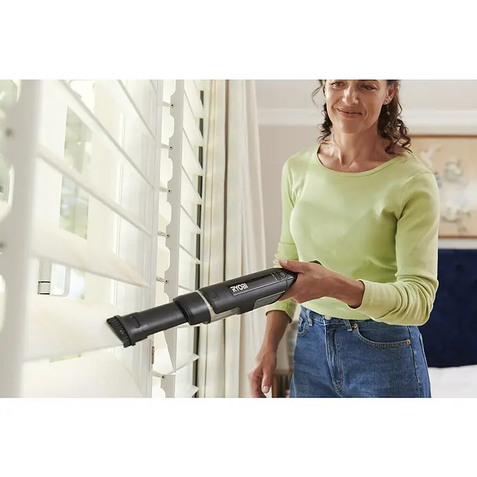 Ryobi Akku-Handstaubsauger RHV44-220G - 8 V, 2 Akkus, 2 Ah, Behältervolumen: 0,15 l - Eine Frau reinigt weiße Fensterjalousien mit einem schwarzen und grauen Ryobi Handstaubsauger.