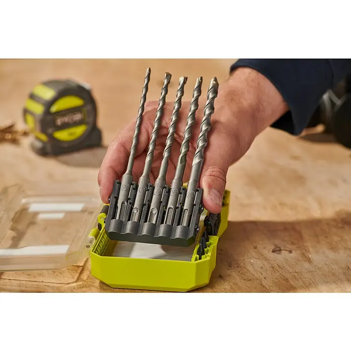 Ryobi Bohrer- & Bit-Set RARXSDS5 - 11 -tlg. - Eine Hand hält ein Set von sechs grauen Bohrern in einem hellgrünen Kasten auf einer Holzwerkbank.