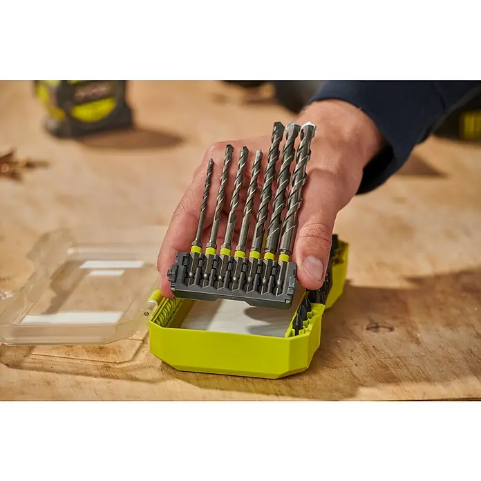 Ryobi Bohrer- & Bit-Set RARXMM7 - 13 -tlg. - Eine Hand hält einen Satz grauer Bohrer mit gelben Ringen in einem neongrünen Koffer auf einem Holztisch.