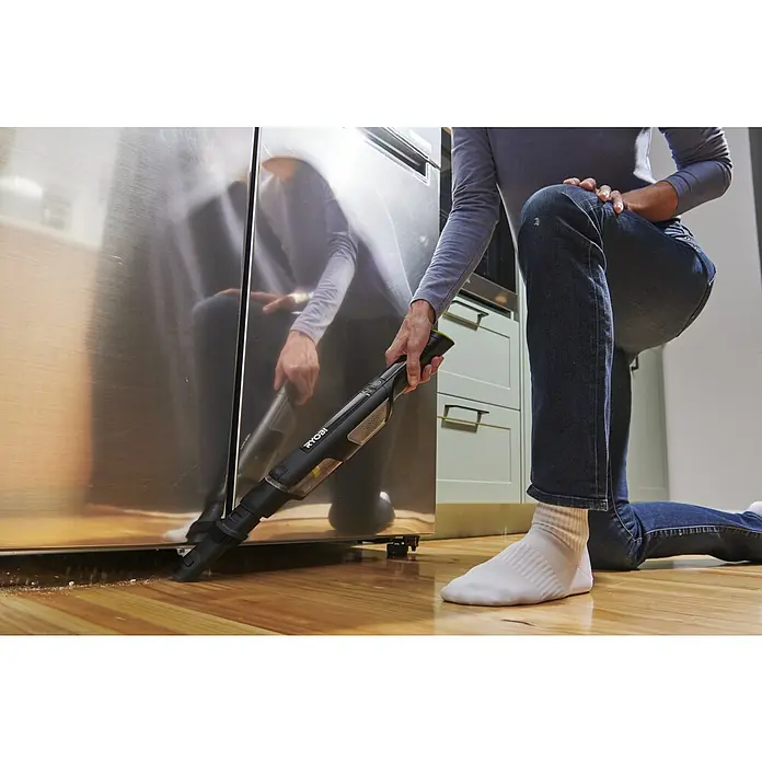 Ryobi Akku-Handstaubsauger RHV44-220G - 8 V, 2 Akkus, 2 Ah, Behältervolumen: 0,15 l - Eine Person kniet und saugt mit einem schwarzen RYOBI Handstaubsauger Krümel unter einem Edelstahlgerät vom Holzboden.