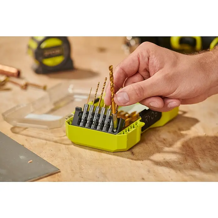 Ryobi Bohrer-Set RARXHG13 - 13 -tlg. - Eine Hand wählt einen goldenen Bohrer aus einem lindgrünen Set. Der Organizer enthält Bohrer verschiedener Größen auf einer Werkbank.