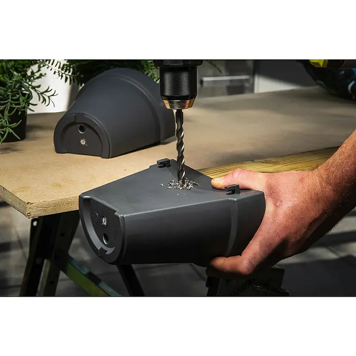Ryobi Bohrer- & Bit-Set RARXMM7 - 13 -tlg. - Dunkelgraues Kunststoff-Pflanzgefäß, in das Drainagelöcher gebohrt werden. Eine Hand hält den Topf beim Bohren.