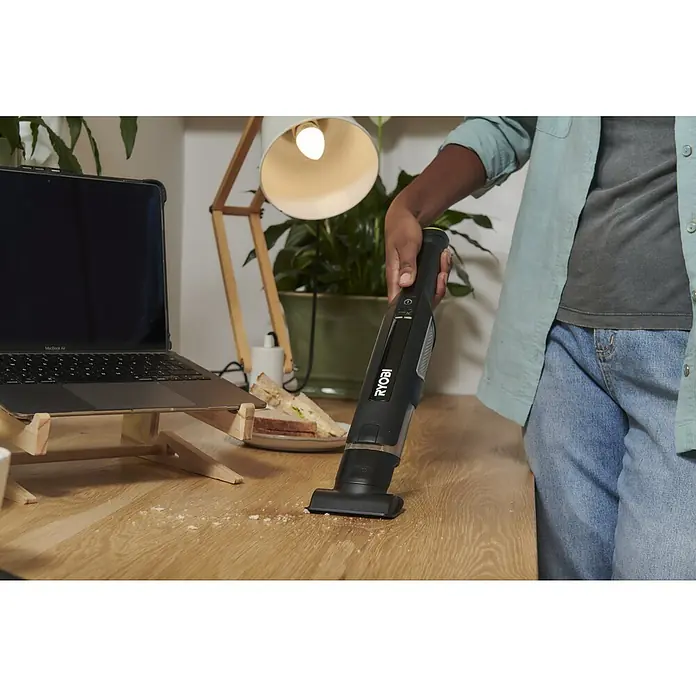 Ryobi Akku-Handstaubsauger RHV44-220G - 8 V, 2 Akkus, 2 Ah, Behältervolumen: 0,15 l - Person reinigt Krümel vom Holzschreibtisch mit schwarzem, gelbem RYOBI Handstaubsauger.