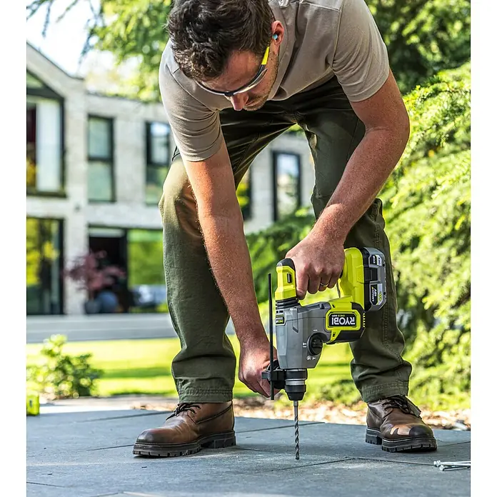 Ryobi Bohrer- & Bit-Set RARXSDS5 - 11 -tlg. - Mann bohrt mit einem gelb-grauen Ryobi Schlagbohrer in eine Terrassenplatte, mit Schutzausrüstung.