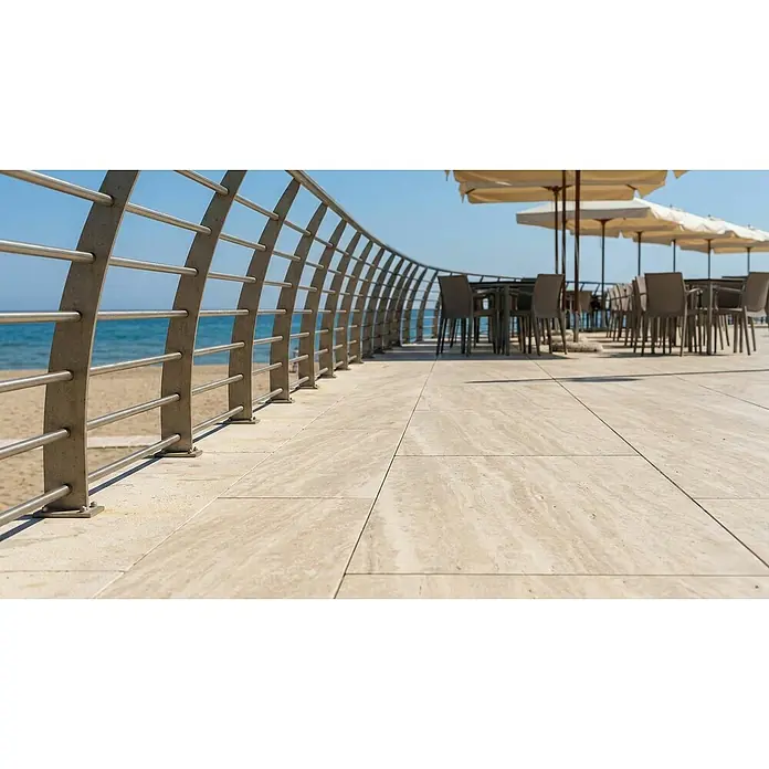 Terrassenfliese Etna - 40 x 80 x 2 cm, Cappucino, Matt - Helle beigefarbene Steinfliesen auf einer Strandcafé-Terrasse am Meer, mit Metallgeländer und verschwommenen Tischen/Sonnenschirmen.
