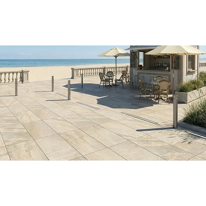 Terrassenfliese Etna - 40 x 80 x 2 cm, Cappucino, Matt - Lineare Entwässerungsrinnen mit silbernen Rosten, eingebettet in helle Terrassenplatten vor einer Strandbar.
