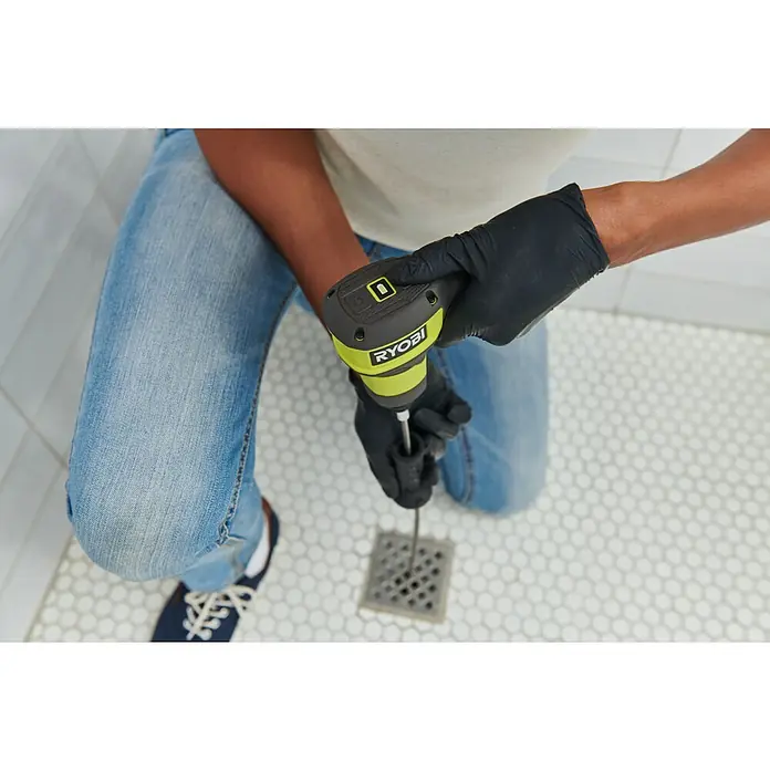 Ryobi 4V USB Akku-Rohrreinigungsgerät RDA4-0 - 4 V, Ohne Akku, Leerlaufdrehzahl: 200 U/min - Person mit schwarzen Handschuhen benutzt ein grün-schwarzes Ryobi Rohrreinigungsgerät, führt dessen Schaft in einen Duschabfluss auf einem sechseckigen Fliesenboden ein.