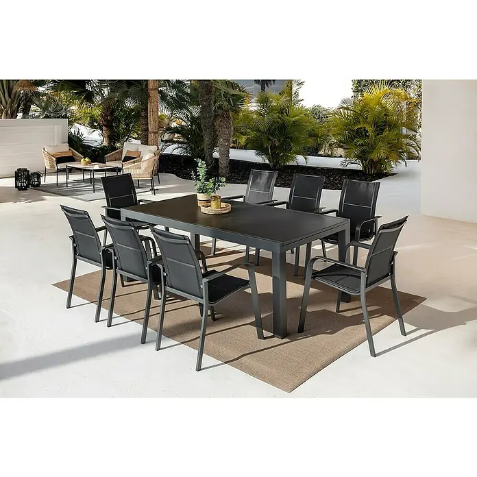 Sunfun Anna Sillón de jardín Anna - 56,5 x 67 x 91,5 cm, Antracita, Acero/Textiline - Conjunto de comedor de exterior con mesa rectangular gris oscuro y ocho sillas a juego, sobre una alfombra de patio entre la vegetación.