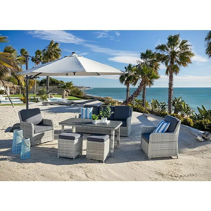 Conjunto de muebles Lodrino - 6 pzs., Aluminio/Olefin, Beige/Gris - Conjunto de salón de exterior de mimbre gris claro con cojines oscuros en un patio arenoso bajo una sombrilla, mar y palmeras al fondo.