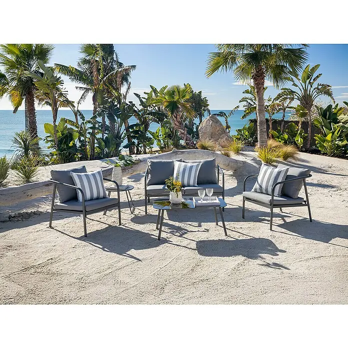 Conjunto de muebles Wesely - 4 pzs., Acero/Poliéster, Gris - Conjunto de salón exterior gris con sofá de dos plazas, dos sillones y dos mesas pequeñas, sobre arena junto al mar.