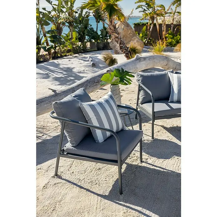 Conjunto de muebles Wesely - 4 pzs., Acero/Poliéster, Gris - Dos sillas de exterior grises con cojines y una mesa pequeña en una playa de arena con palmeras y el océano de fondo.